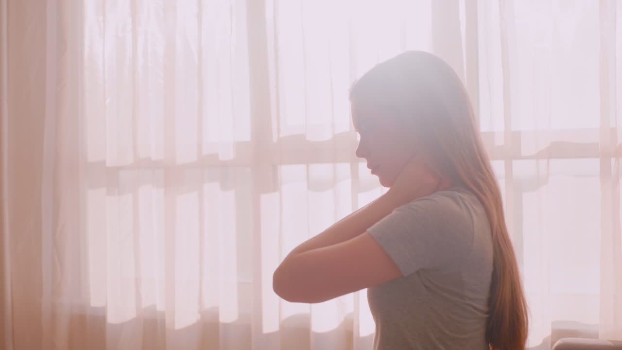 Side view of independent girl sitting beside bright sunlit window gently stretching neck with both hands due to discomfort or stiffness, showing calm expression in soft natural light
