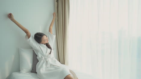 아시아 여성은 침대에 누워서 흰색 드레싱 가운을 입고 머리 위로 팔을 뻗고, 잠에서 깬 후 아침 루틴을 하고 있습니다.