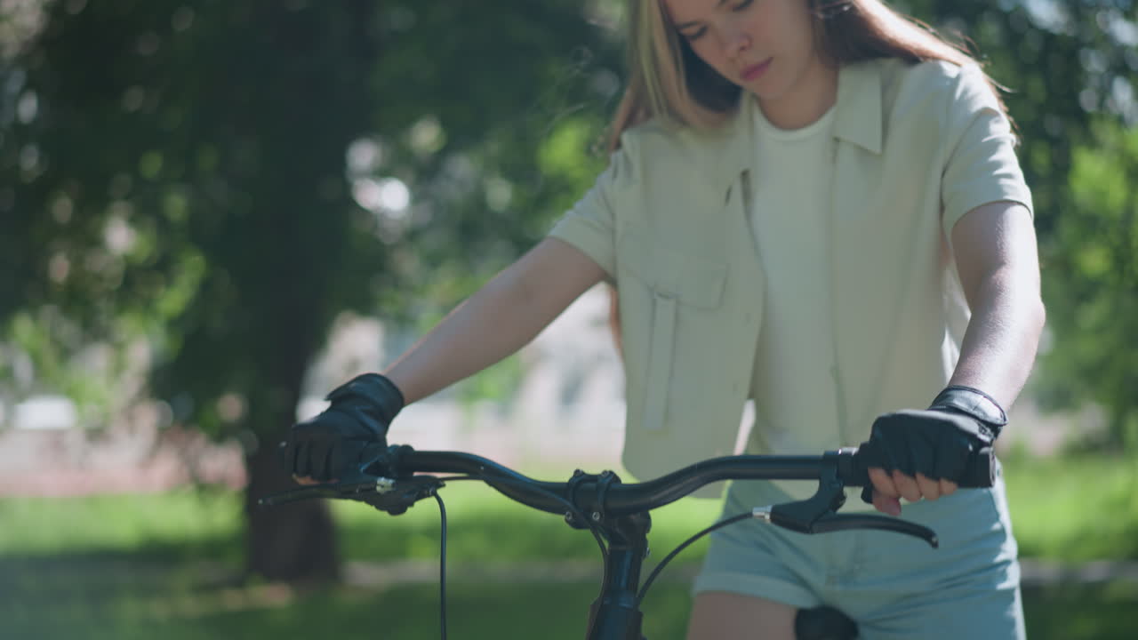 검은색 자전거의 손잡이를 잡고 있는 여성의 클로즈업, 자전거 장갑을 입고, 배경에는 잎이 부드럽게 바람에 움직이는 울창한 초록색이 있습니다.