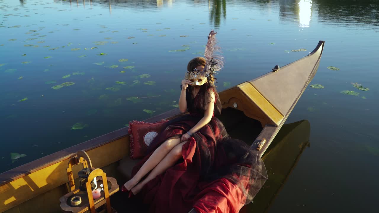 Gorgeous model in luxury dress with bare legs sitting in a boat and holding mask on a face at sunset. Natural background of evening and a woman in carnival clothes floating on the river.