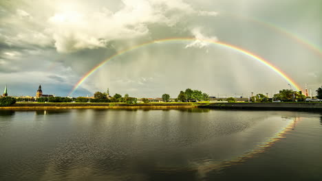 Aufnahme-Von-Weißen-Wolken,-Die-Im-Zeitraffer-Durch-Einen-Regenbogen-Vorbeiziehen,-Der-Sich-Nach-Regenfällen-über-Dem-See-Bildet,-Mit-Blick-Auf-Die-Stadt-In-Der-Ferne