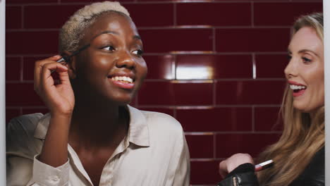 Two-Young-Female-Friends-Doing-Make-Up-Reflected-In-Mirror-Of-Bathroom-In-Club