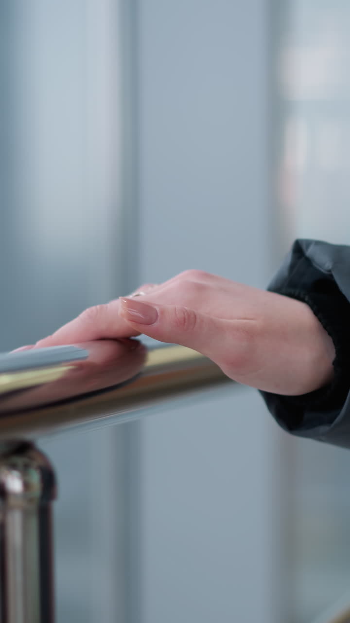 primer plano de la mano de la mujer con anillo deslizándose en el carril brillante mientras camina a través de un edificio moderno con reflejos de vidrio y luces parpadeantes en el fondo