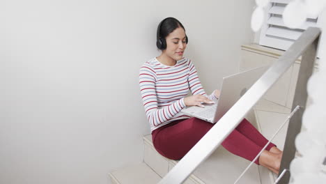 Happy-biracial-woman-sitting-on-stairs-using-headphones-and-laptop,-slow-motion