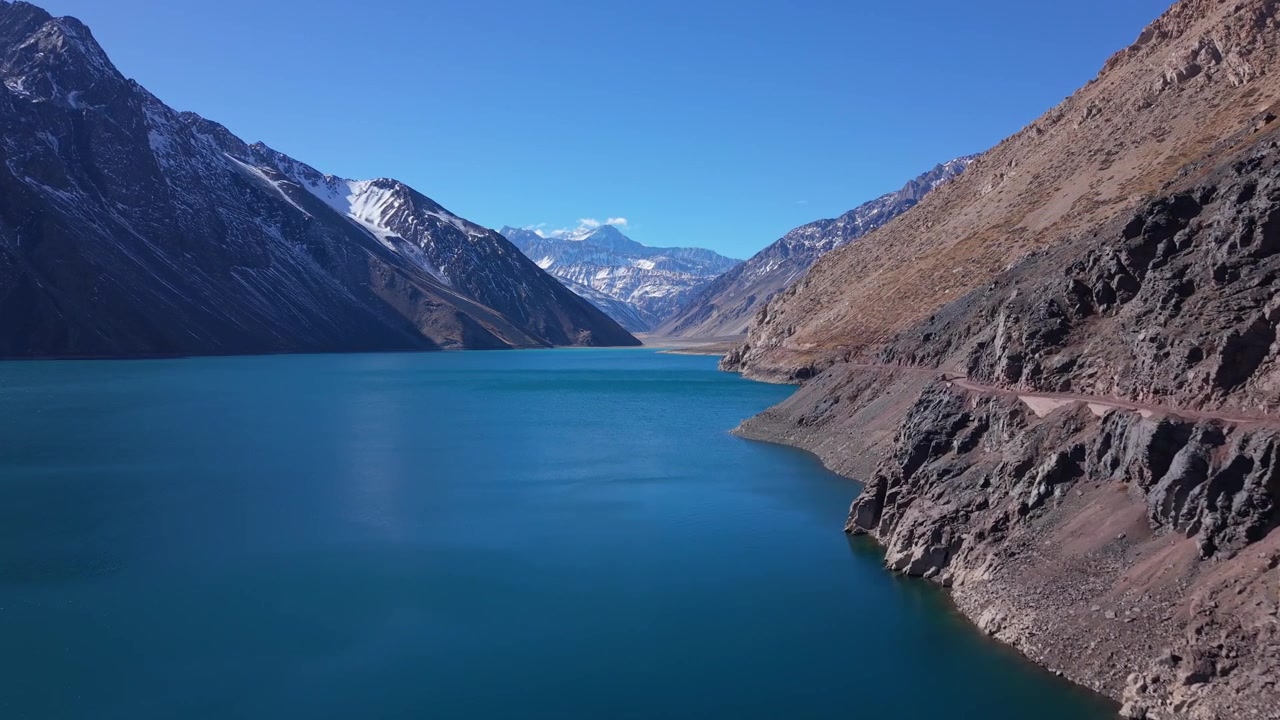 Volo planato sopra le acque turchesi di un lago glaciale che rivela cime andine innevate che si ergono sopra la costa rocciosa arida. Embalse del Yeso, Cile.