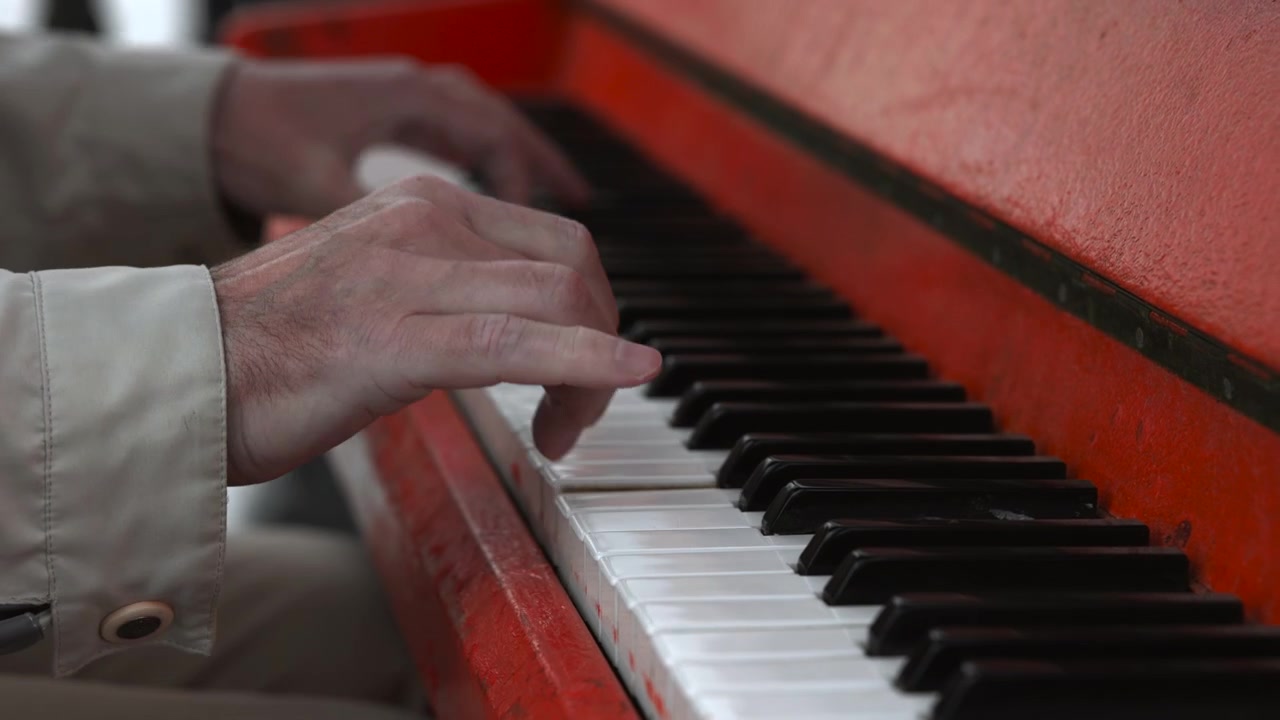 En pianist er dybt koncentreret om at spille klaver i et indendørs studie. Nærbilledet fremhæver hænderne og tangenterne, og viser de sorte og hvide tangenter samt det røde klaverhus