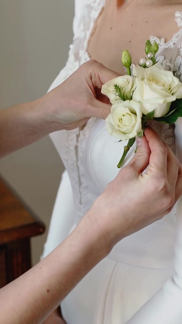 Vertical video: Offering flowers, helper pinning white rose to left lace bodice in bride prep room
