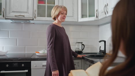 close-up rear view of woman reading novel at home while her mother walks into the kitchen, looking at her with a warm smile, creating a cozy and intimate family moment in a modern home setting