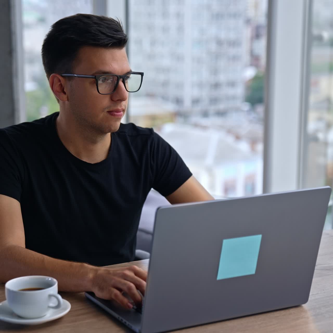 Pensive young man in glasses and black t-shirt sits in the office in front of laptop. Male looks into window thinking over something. Blurred backdrop