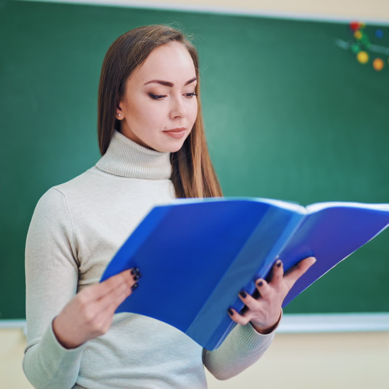Young teacher with a folder in the classroom. Attractive pedagogue holds a blue folder and looks for the necessary information. Educational concept.