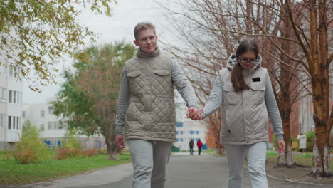 happy moment of young couple walking hand in hand, swinging arms joyfully in sync while smiling at each other on peaceful park pathway with blurred people in background