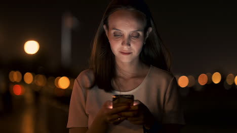 at night a young girl sits on the grass in a big city and looks into the smartphone screen writes a message and maintains her personal blog.