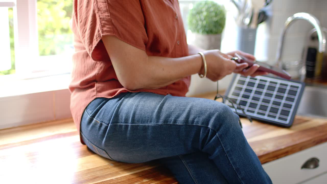 Ældre asiatisk kvinde i rustfarvet top på bænk trykker på telefon, ser kabel tilsluttet solpanel, der oplader
