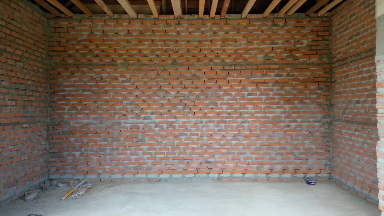 Un paneo lento hacia la derecha recorre un interior vacío y sin terminar con paredes de ladrillo rojo, mortero gris, vigas de madera expuestas, materiales de construcción dispersos y una suave luz ambiental