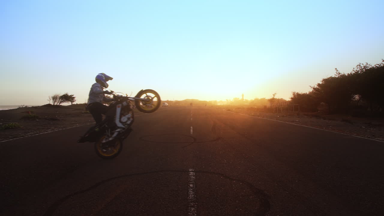 A motorbike stunt rider performs circle wheelies while standing up on his motorcycle. Shot on the back roads of Indonesia at sunset.
