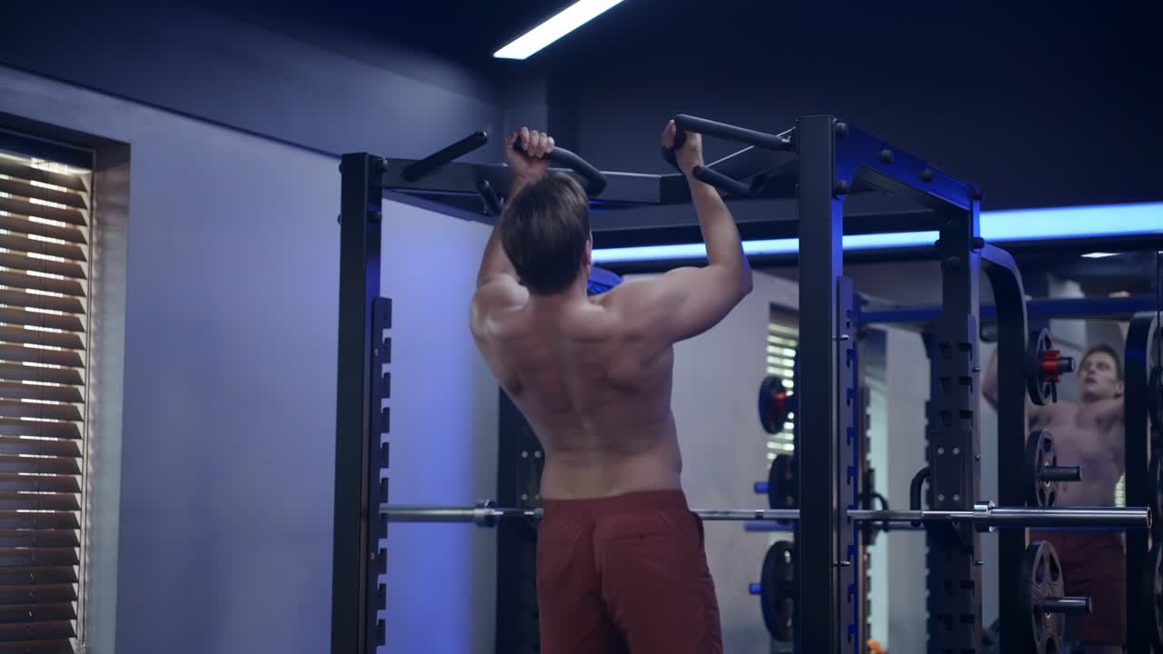 Shirtless blond fit man doing pull ups at dim lit modern gym - rear view