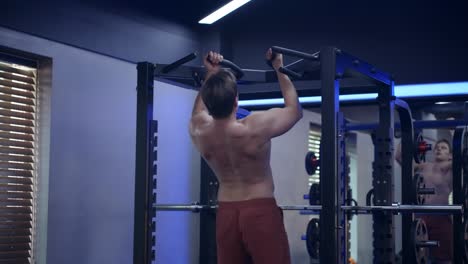 shirtless blond fit man doing pull ups at dim lit modern gym - rear view