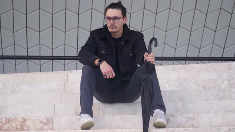 한 젊은이가 계단에 앉아 슬프고 좌절한 모습으로 검은색 재과 안경을 입고 우산을 쓰고 있다.