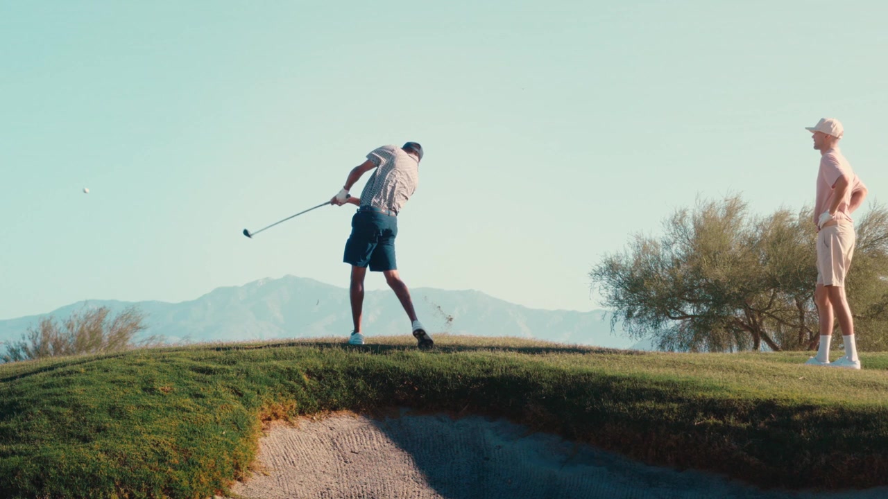 Two golfers stand on a grassy ridge as one hits wood off a hill against a desert mountain range at golden hour in slow motion, a cinematic sequence for lifestyle and travel edits.
