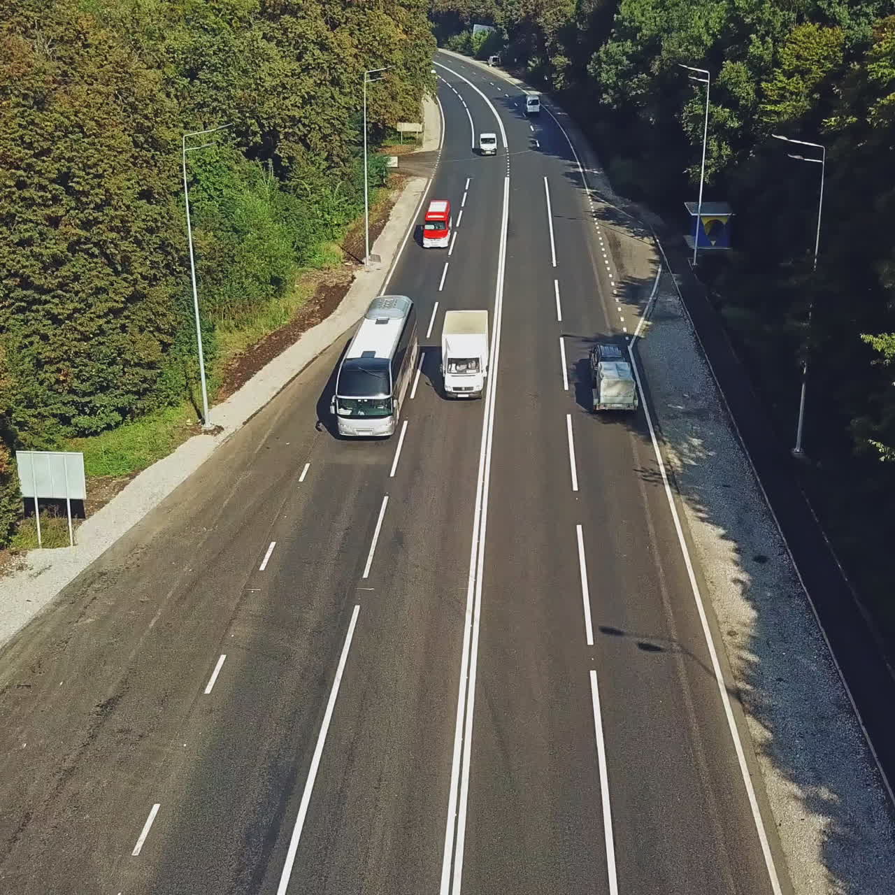 New road with four lines and the cars going by in summer. Green trees growing on roadside of a two-lane road. Bus stop from the right side of the road. Aerial view along the highway