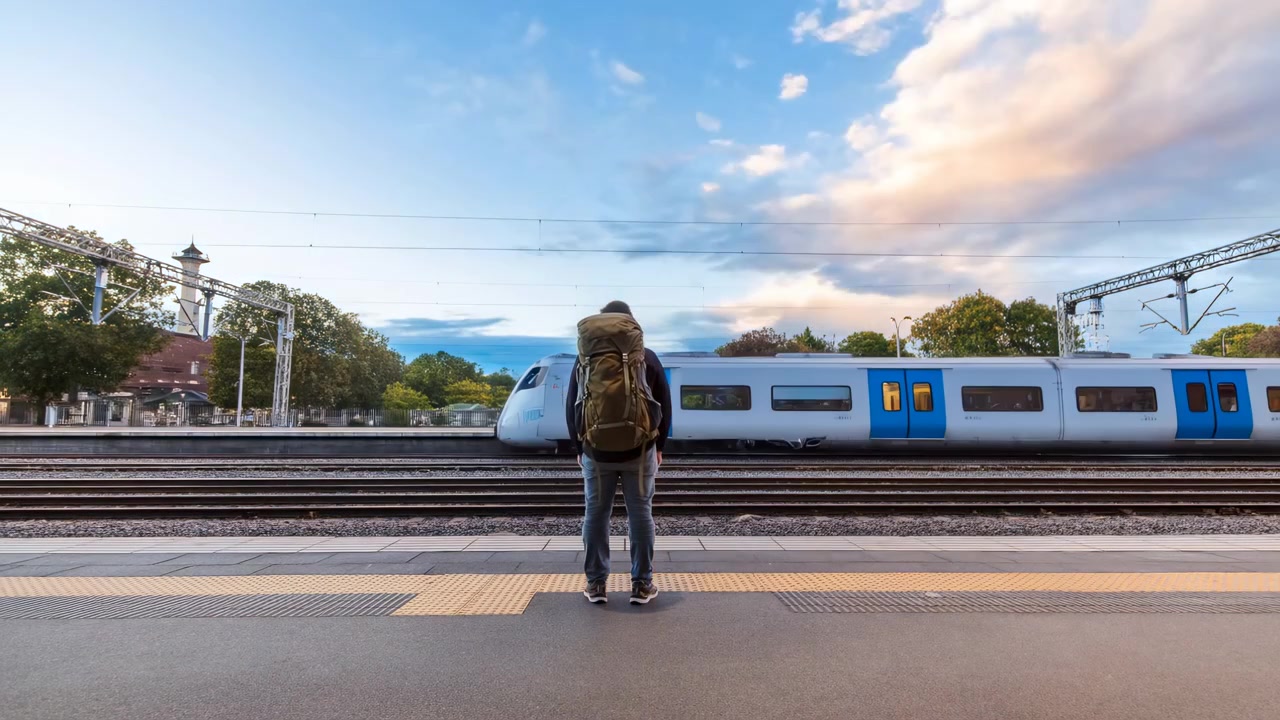 Rear view of lone backpacker standing on railway platform while commuter train rushes by as wide horizontal blur, warm open doorway light glowing against cool dusk station setting, with backpacker