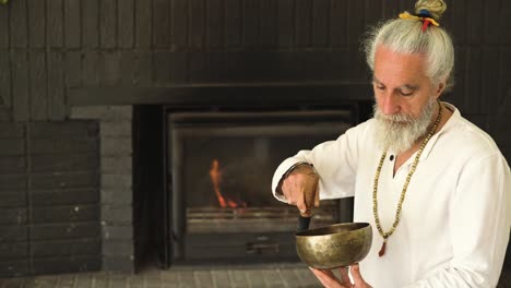 senior man playing bowl gong with mallet against fireplace