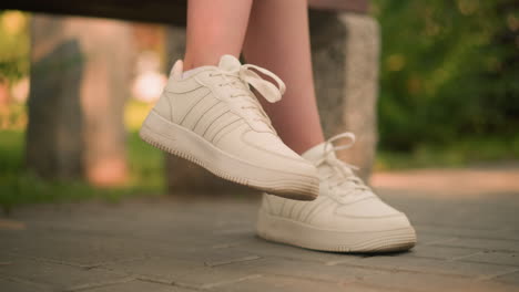 white person seated outdoors with one leg slightly lifted wearing white sneakers, gentle movement of leg with blurred background softly illuminated by sunlight