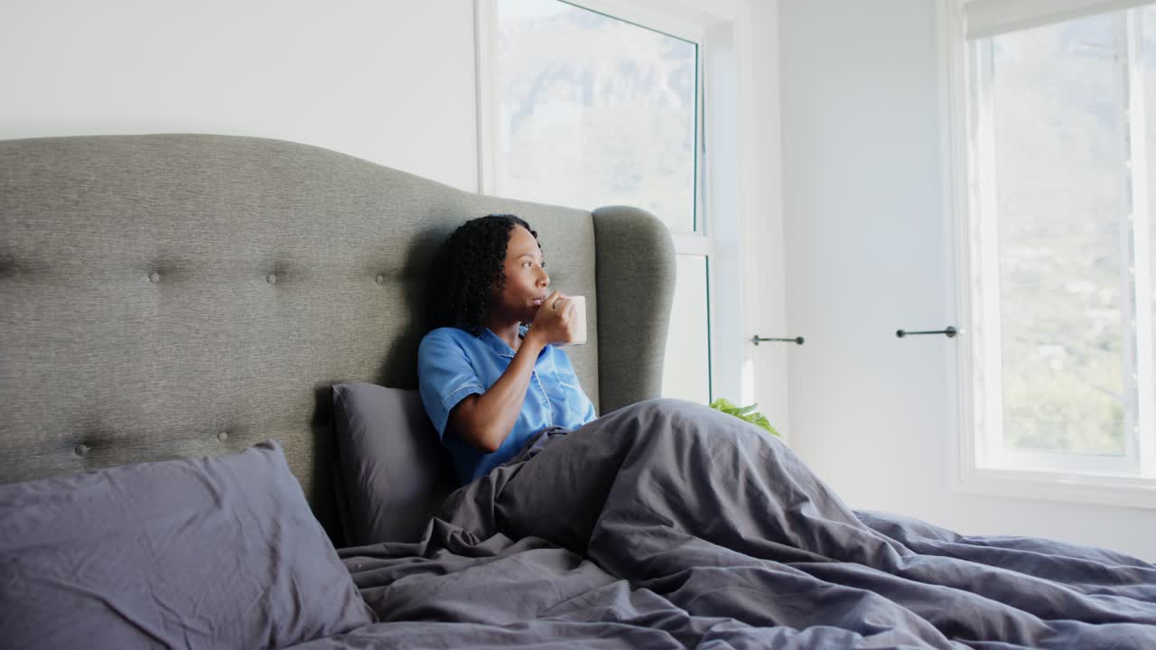 Donna afroamericana adulta seduta sul letto, che solleva una tazza, sorseggia, guarda fuori dalla finestra, con spazio per il testo