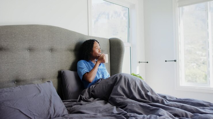 Voksen afroamerikansk kvinde sidder på sengen, løfter krus, nipper til drik, kigger ud af vinduet, med plads til tekst.