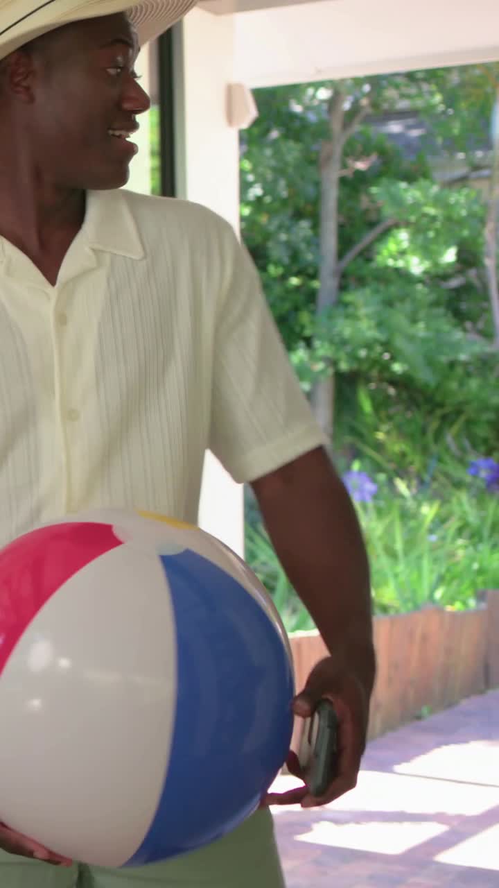 Vertikal video: Dörren öppnas, man i hatt kliver in i hemmet för att möta vänner och håller i en badboll.