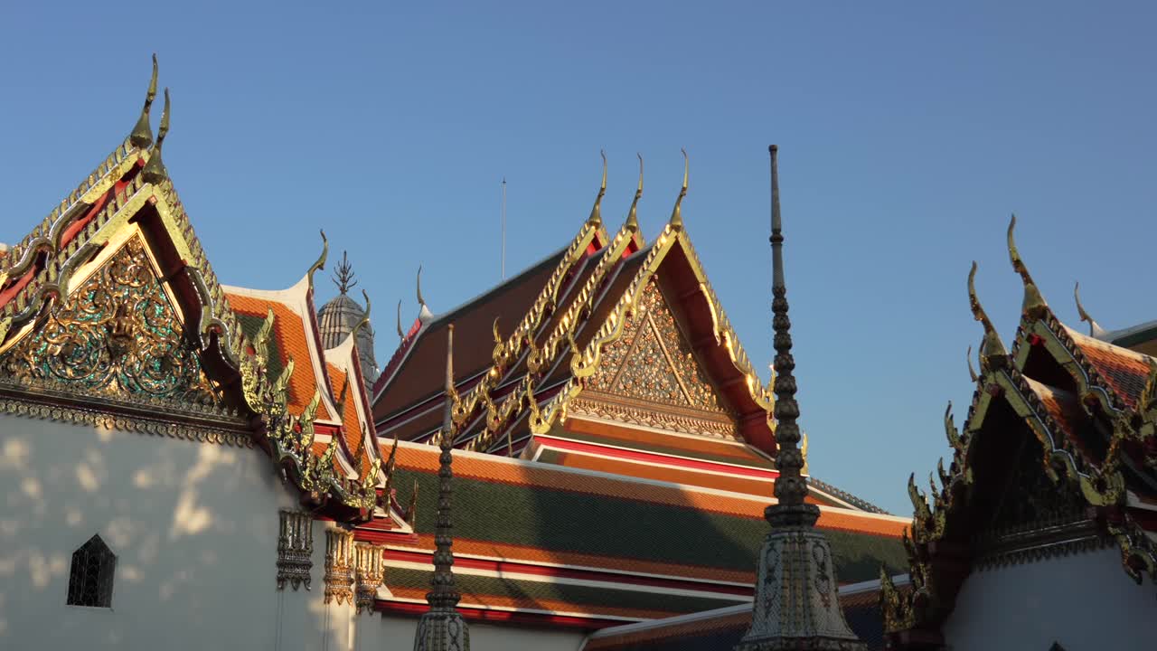 Il complesso del tempio Wat Pho mostra diversi tetti con intricati timpani dorati, piastrelle colorate ed elementi decorativi sotto un cielo azzurro a Bangkok, al rallentatore