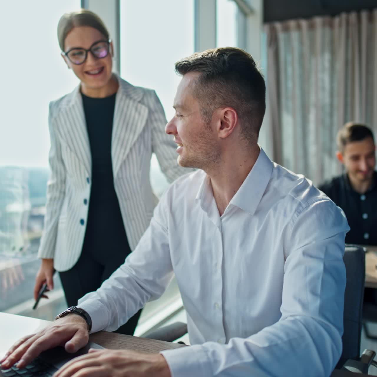 Cheerful smiling colleagues talking in the office. Female stands near her male coworker discussing something and laughing. Male employee sits at desk at backdrop