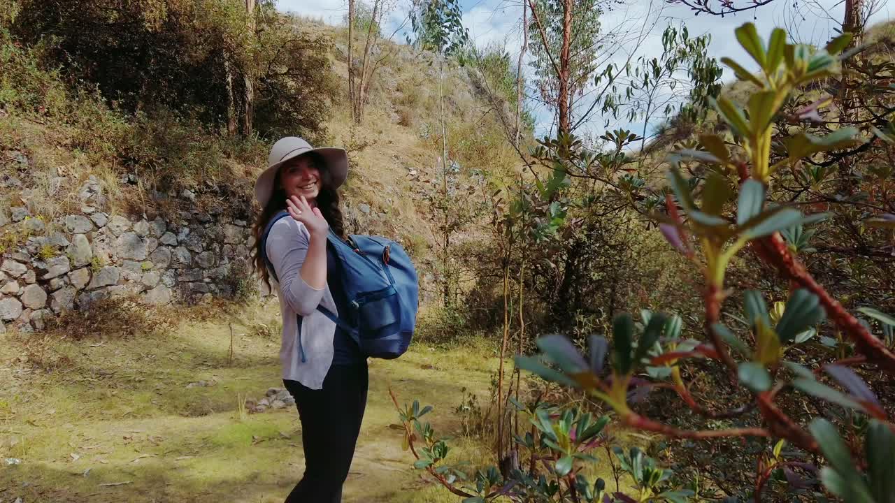 Reveal shot of a woman traveler with long brown hair, smiling at waving at the camera while on an old Inca trail near Cusco, Peru.