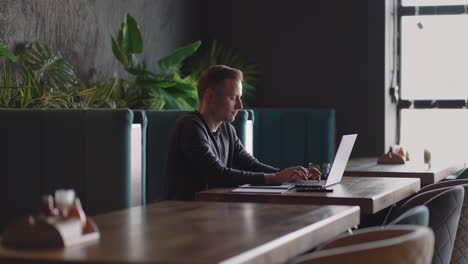 A-young-man-in-a-shirt-is-sitting-at-a-table-with-a-laptop-and-typing-on-the-keyboard.-A-student-can-study-remotely.-A-businessman-conducts-his-business-remotely