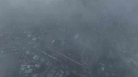 Aerial-drone-top-down-shot-through-clouds-over-an-old-fortress-wall-surrounded-by-Umerkot-city,-Tharparkar,-Pakistan-on-a-cloudy-evening