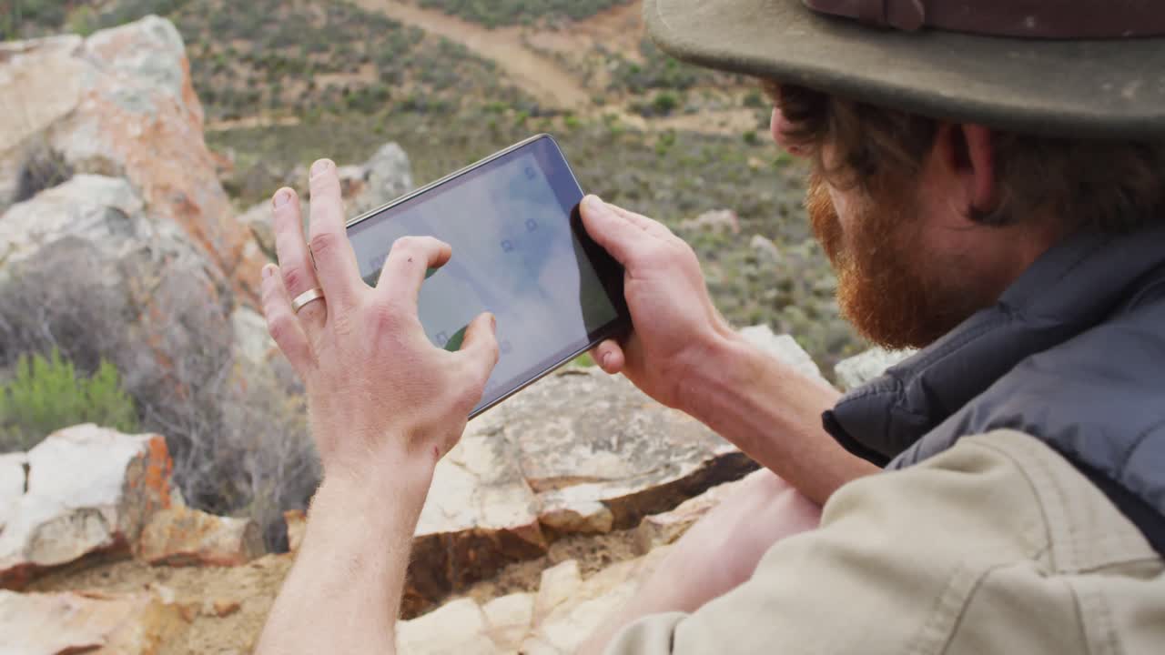 Bearded caucasian male survivalist sitting on mountain, using tablet