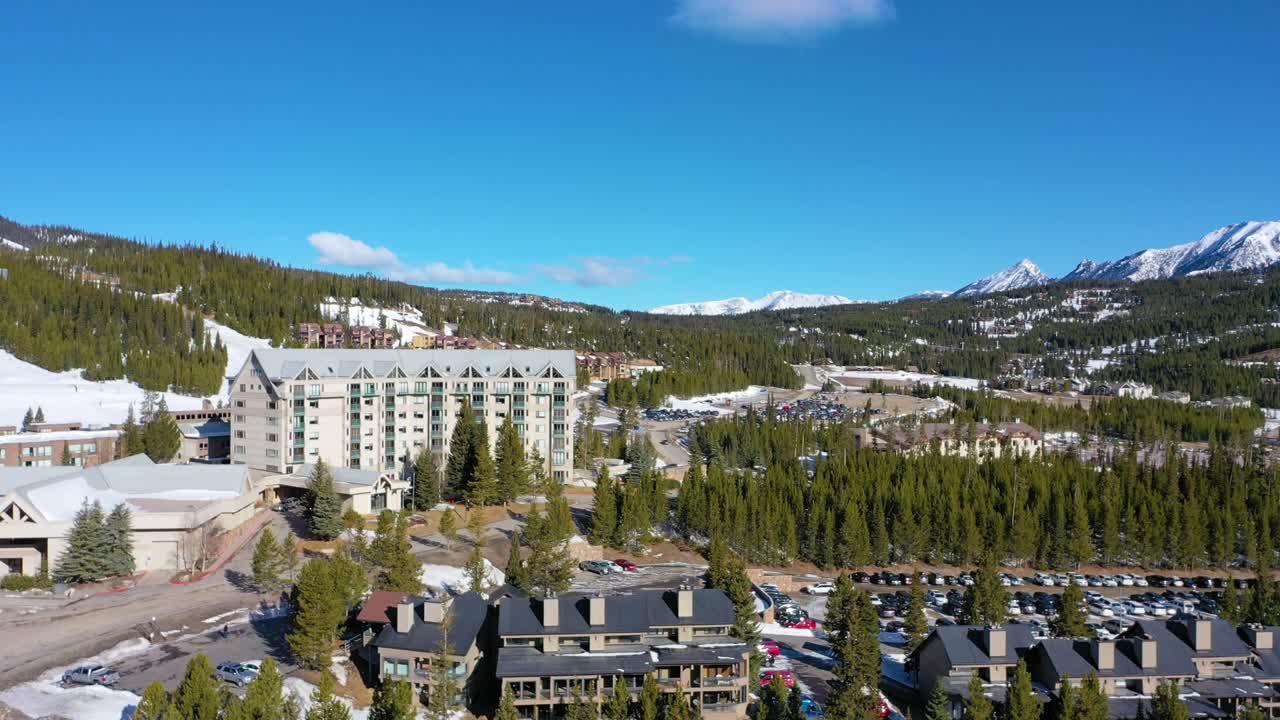 Aerial drone footage captures a packed parking lot in snowy Big Sky, Montana, just below the ski resort, with surrounding vacation condos, tall pines, and vast mountain scenery all around