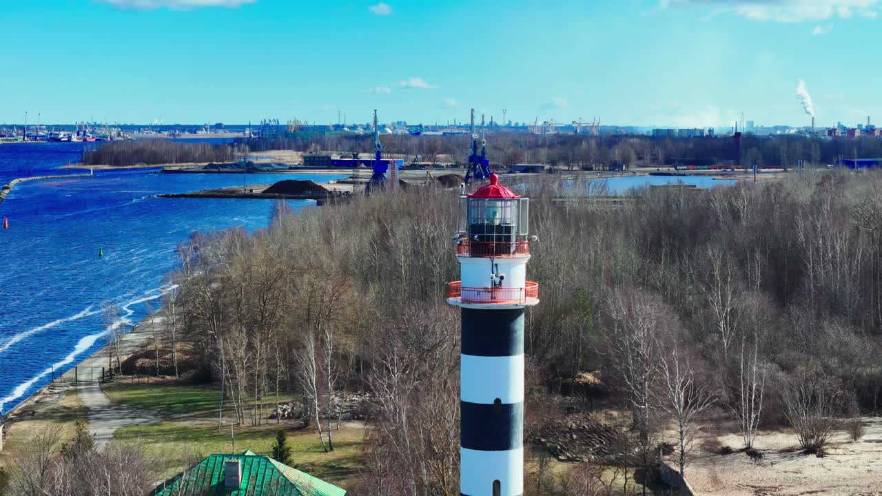 A striped lighthouse stands on the riverbank near leafless trees and industrial cranes, marking the entrance to a port where land meets water under a clear sky.