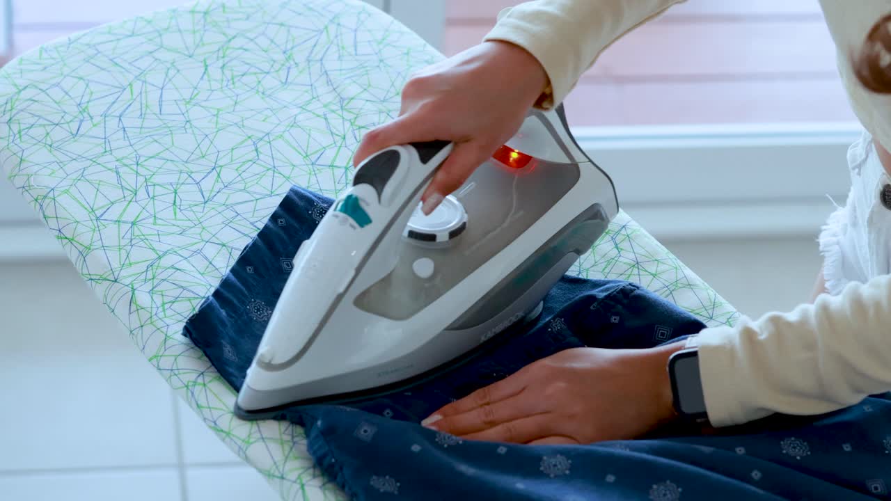 A woman uses a steam iron to press a blue patterned shirt on an ironing board in a bright, sunlit room with soft natural light