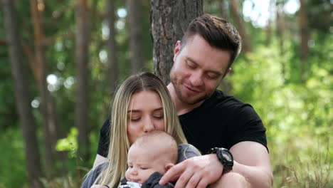 caucasian family of three sitting in the woods. woman kisses her baby to a head.