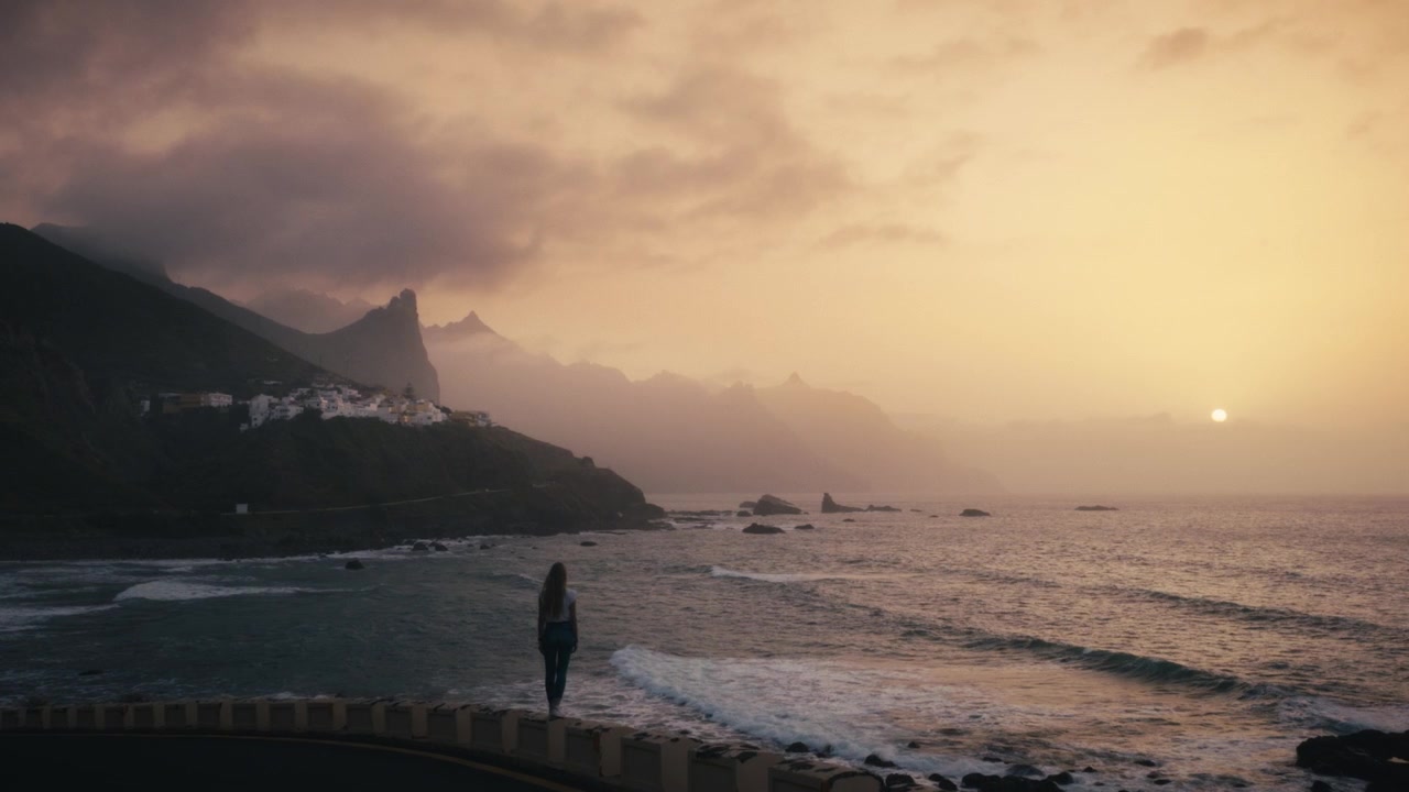 An aerial view of a woman silhouetted against the setting sun on a coastal barrier near Benijo village in the Anaga mountain range, Tenerife, Spain, with golden light over the clouds and sea.