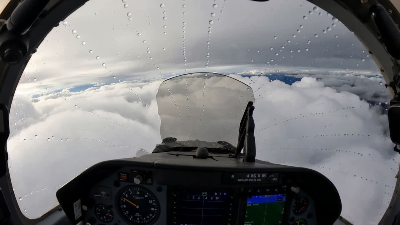 Telecamera della cabina di pilotaggio di un aereo che riprende il velivolo che si inclina in una nuvola umida, con goccioline che si formano sul parabrezza.