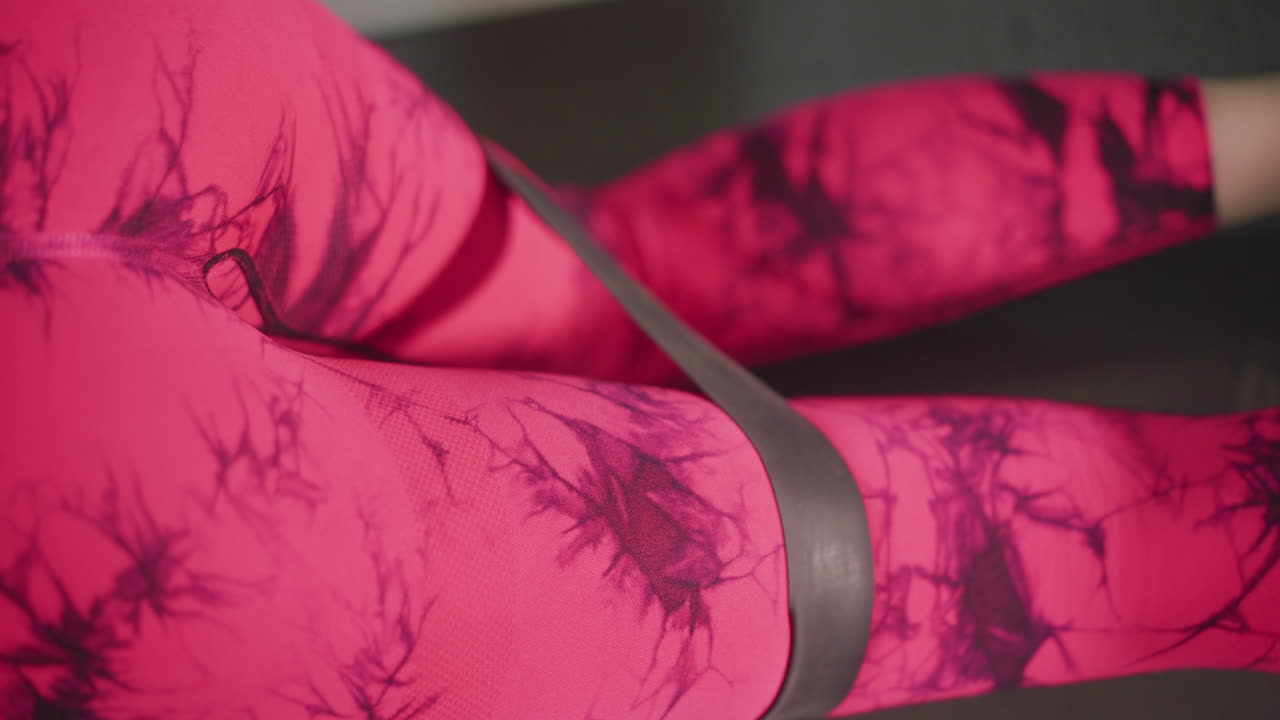 Primo piano: leggings rosa durante un esercizio di compressione dei glutei con fascia elastica su tappetino, tecnica riabilitativa guidata dal terapista, contrazione muscolare controllata, dettaglio del tessuto texturizzato, illuminazione da studio clinico, focus sul movimento preciso.