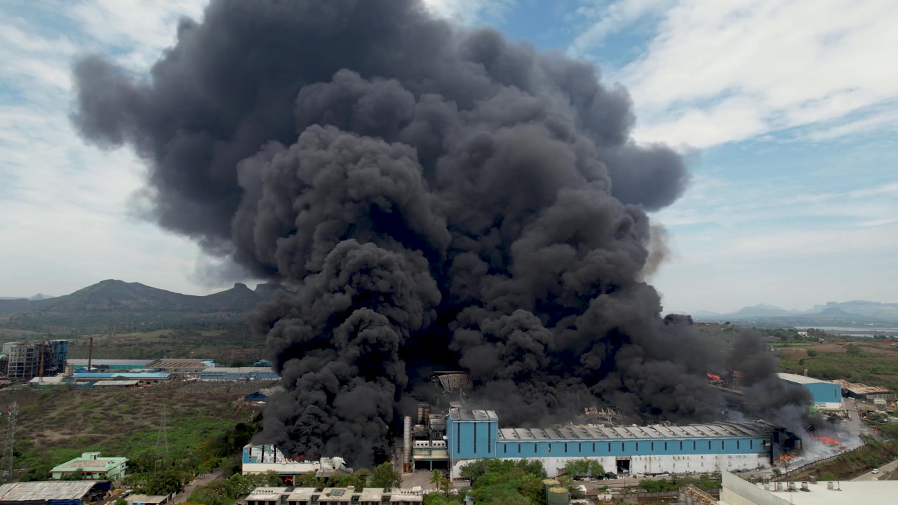 Primer plano aéreo con dron de un incendio masivo en una fábrica que produce denso humo negro sobre un área industrial con colinas y edificios circundantes.