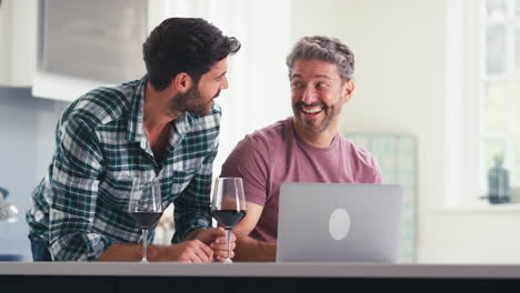 Same-Sex-Male-Couple-Drinking-Wine-Using-Laptop-At-Home-To-Check-Finances-Book-Holiday-Or-Shop