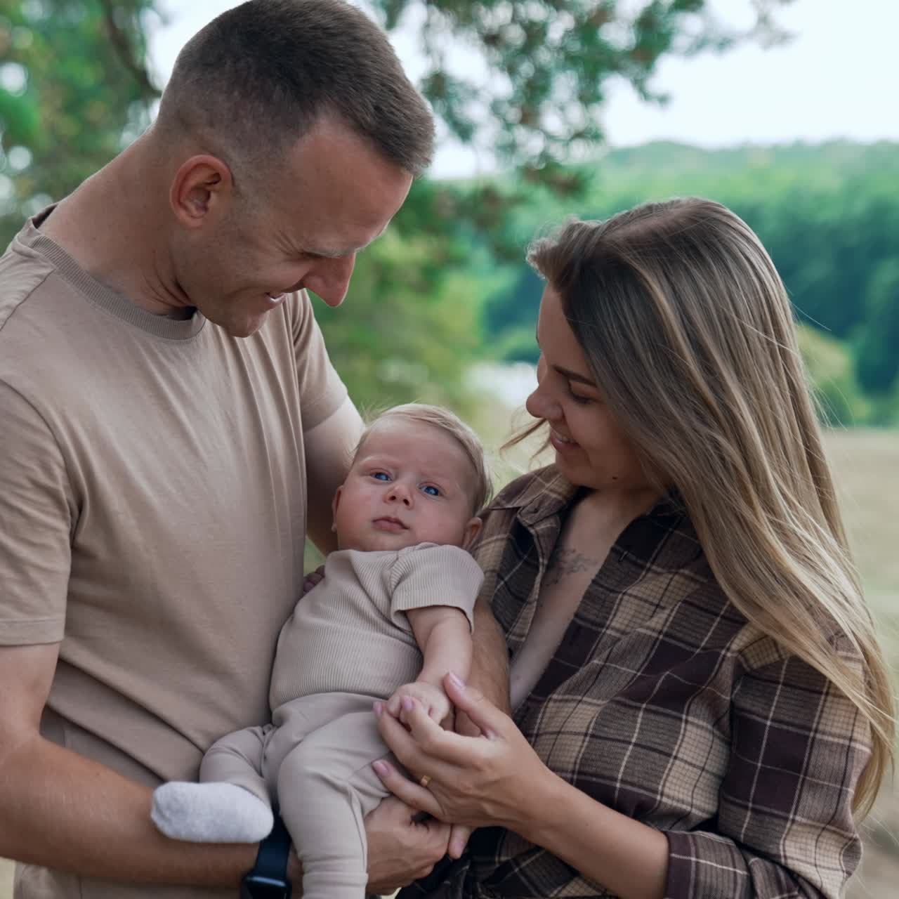 Una coppia felice è in piedi nella natura e tiene un bambino tra loro. I genitori guardano il loro neonato con amore e poi si baciano.