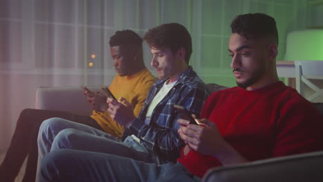 three friends using smartphones on a couch at night