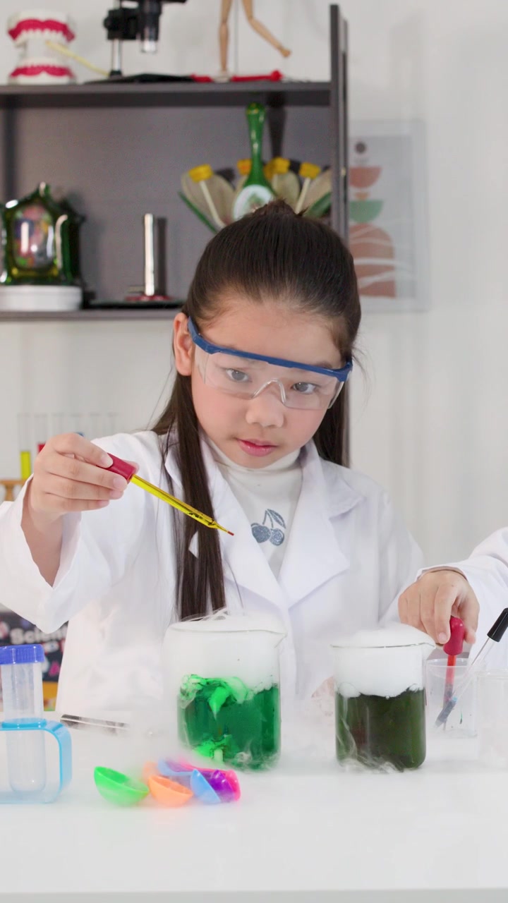Seorang gadis muda mengenakan kacamata pelindung menggunakan pipet untuk eksperimen kimia yang berwarna-warni