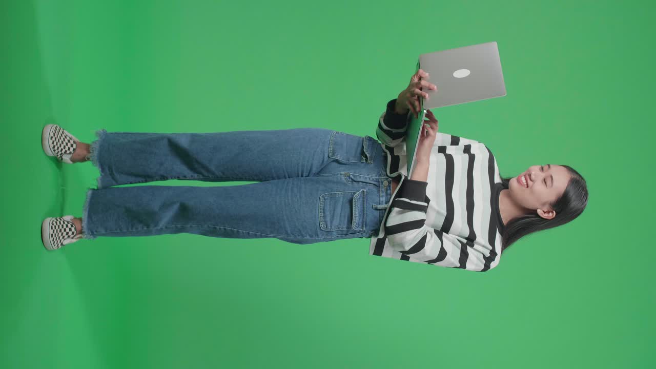 cuerpo lleno de una mujer asiática sonriente diciendo wow mientras usa la computadora en el fondo de la pantalla verde