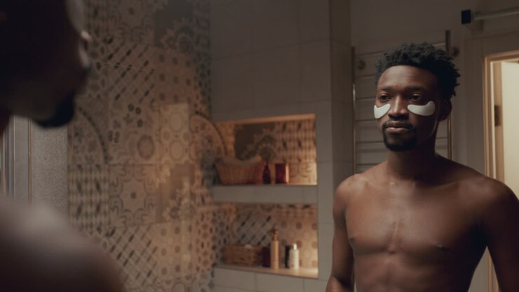 Uomo afroamericano che applica patch sotto gli occhi davanti allo specchio del bagno.